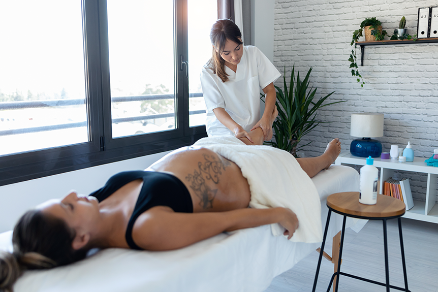 Physiotherapist providing a gentle foot and leg massage to a pregnant woman lying on a treatment table in a bright, calming clinic.