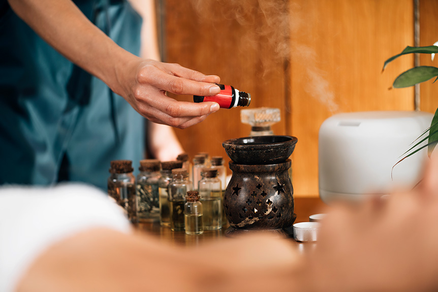 Therapist pouring aromatic essential oil into a diffuser as part of an aromatherapy massage setup to enhance relaxation and sensory experience.