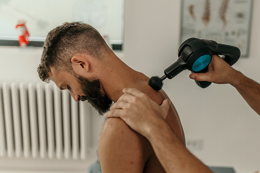 Physiotherapist applying a massage gun to the neck and shoulder muscles to ease deep tissue tightness.
