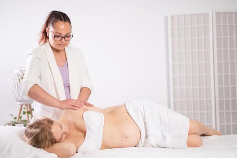 Ethnic masseuse massaging resting young pregnant woman on bed in parlor