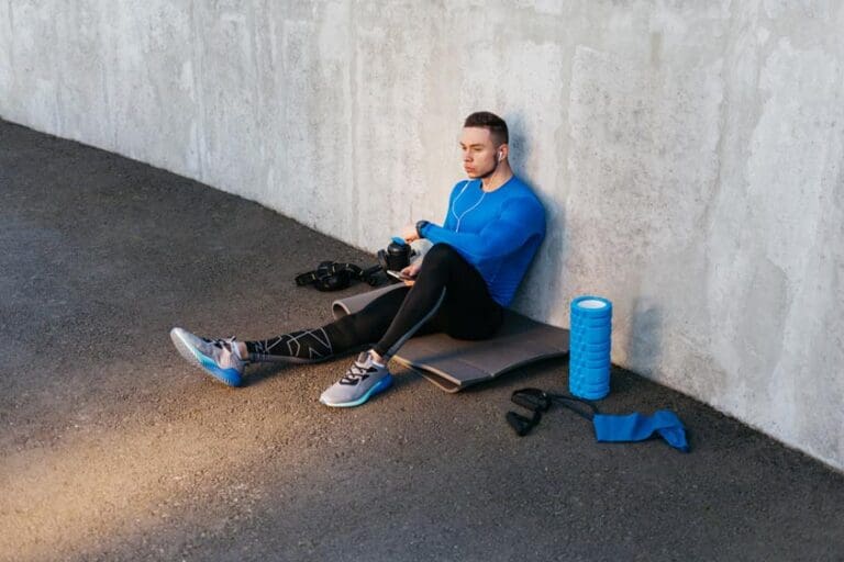 Young Sports Man In Blue T-Shirt Siting On Mat And Listening Music On The Background Of Gray Wall