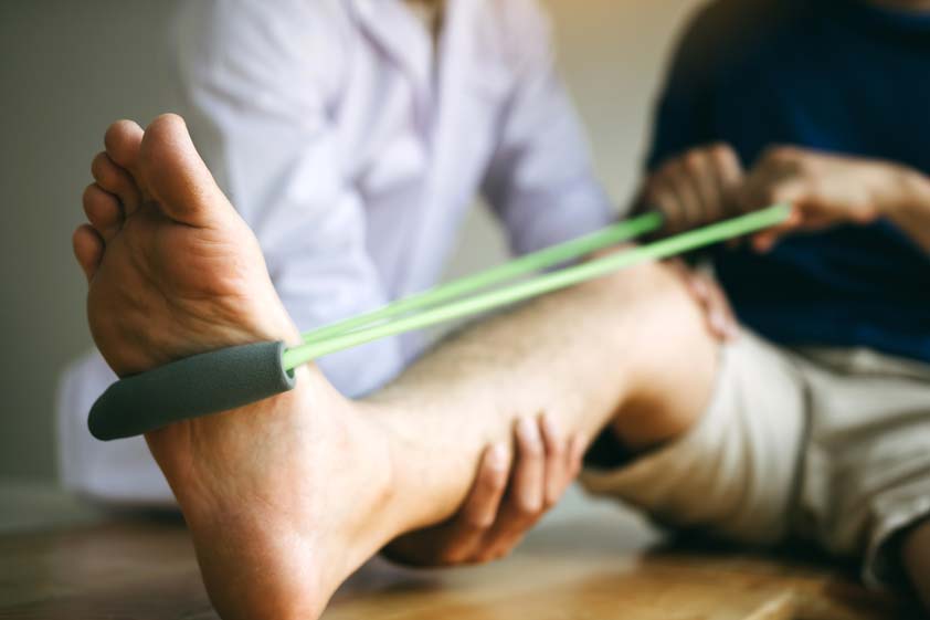Patient use resistance band stretching out his leg with physical therapist helps in clinic room.